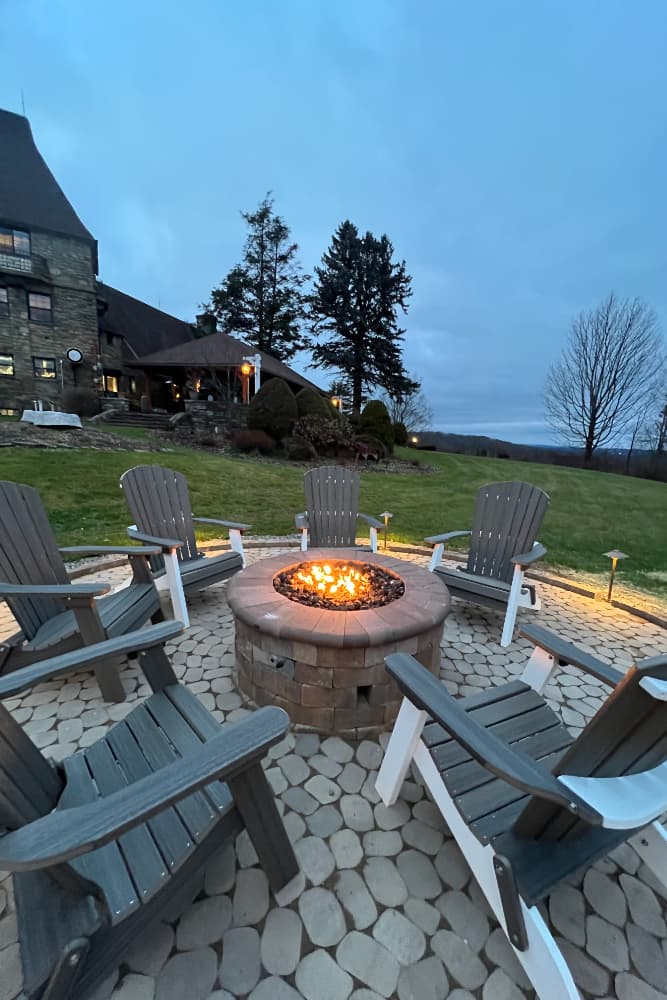 A cozy outdoor fire pit surrounded by Adirondack chairs on a stone patio. A large stone building with a dark roof and illuminated windows is visible in the background. Tall trees and a clear sky complete the scene.