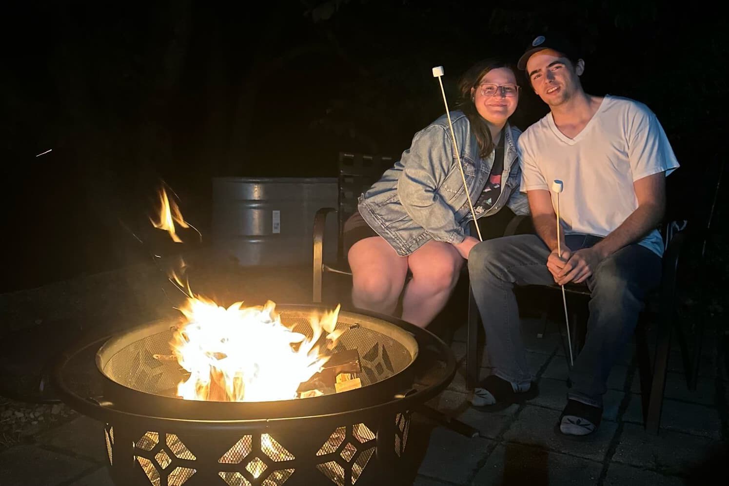 A couple sits by a fire pit at night, roasting marshmallows on sticks.