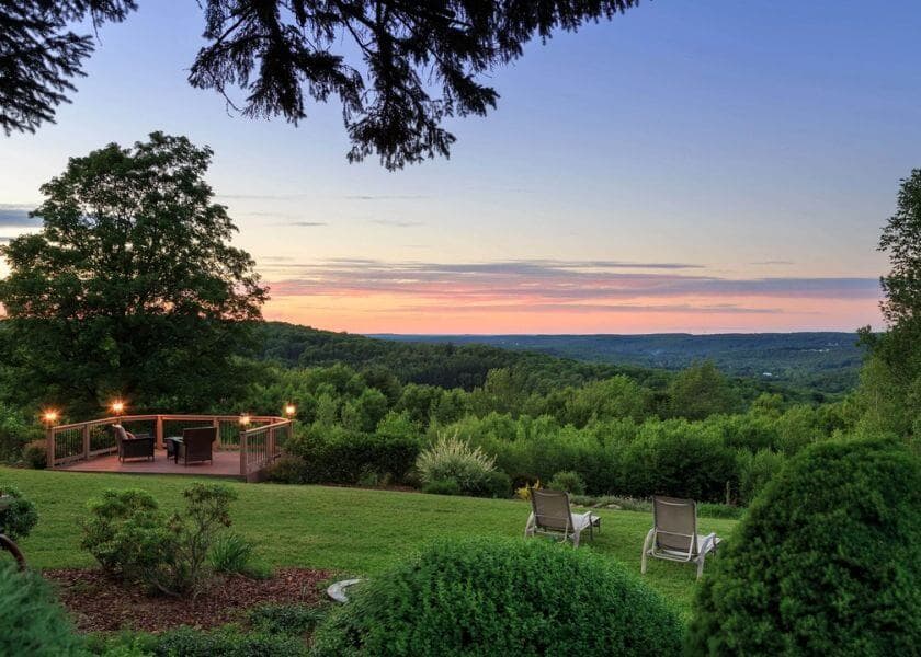 A serene landscape featuring lush greenery, a wooden deck, and two lounge chairs under a colorful sunset sky.