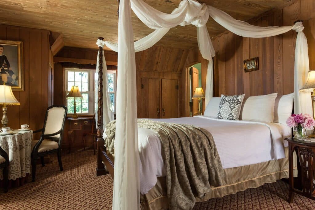 A cozy, warmly lit bedroom featuring a canopied bed, elegant decor, and a small seating area by a window.