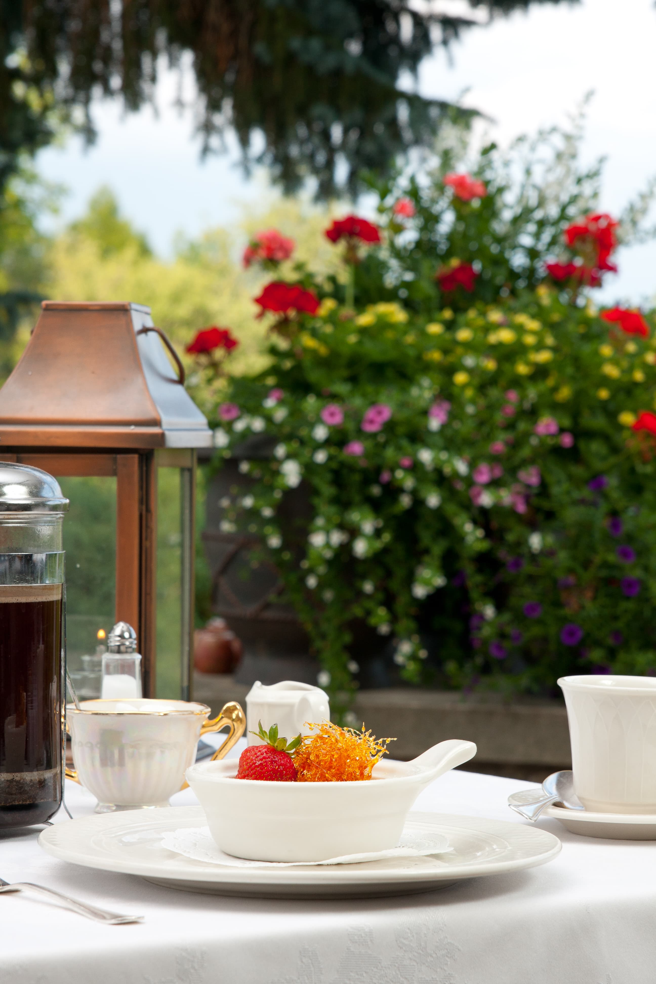 A table setting outdoors with a white plate holding a bowl of fruit and a cup of coffee. A lantern and a sugar bowl are also on the table. In the background, there is a lush garden with colorful flowers.