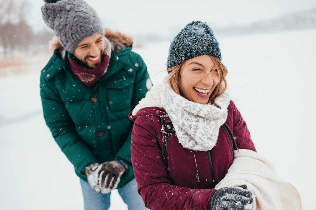 A couple enjoys a snowy day, laughing and bundled in winter clothing.