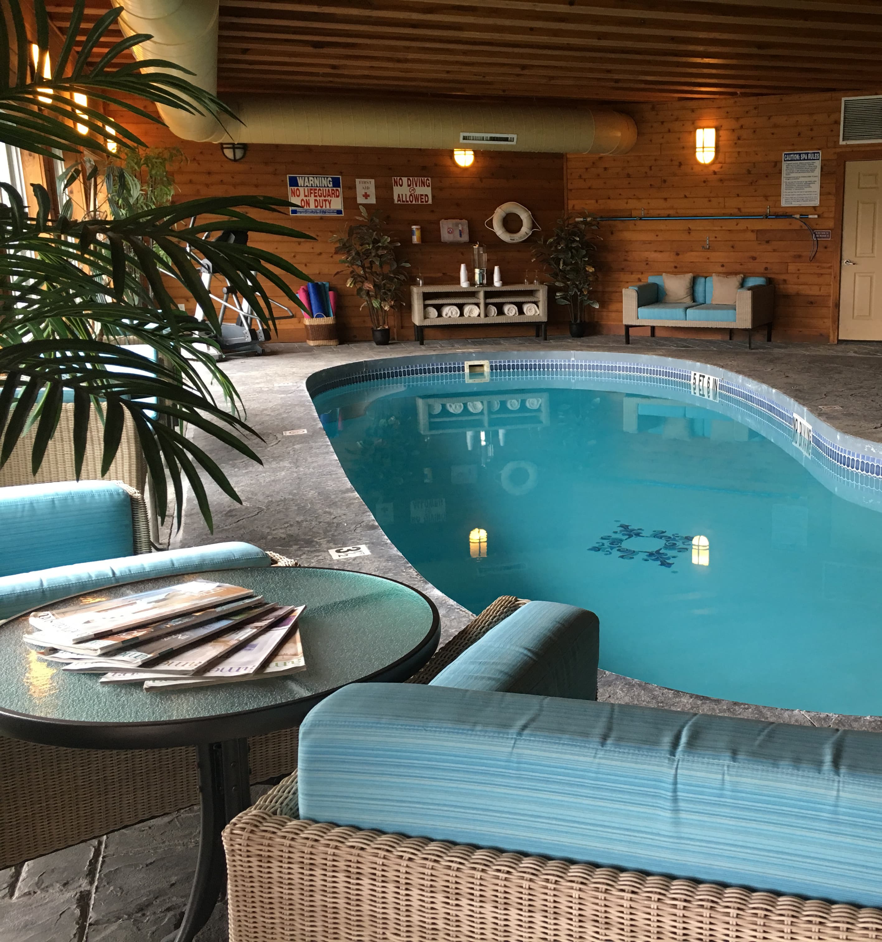 An indoor pool with a curved shape and clear blue water. The pool area is surrounded by comfortable seating, including armchairs and a small table with magazines. There are warning signs about no diving and no lifeguard on duty, as well as a life ring and other pool safety equipment. The ceiling is high with exposed wooden beams.