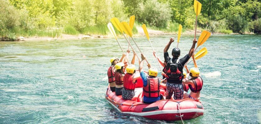 A group of people in life jackets paddle a red raft on a river, celebrating with raised arms.