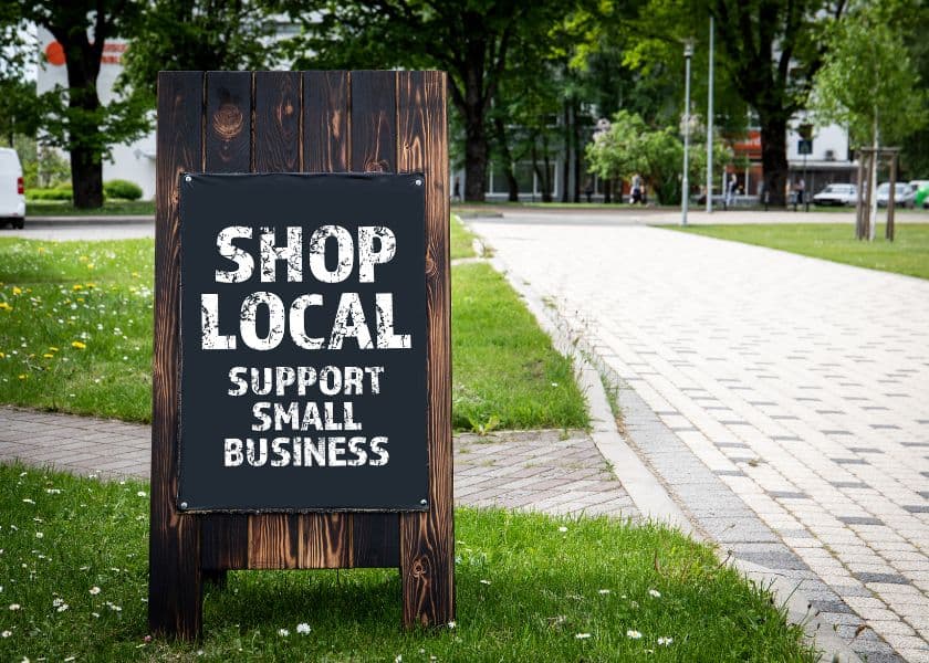 wooden sidewalk sign reading shop local support small business along a tree-lined street with grass and paved walkway