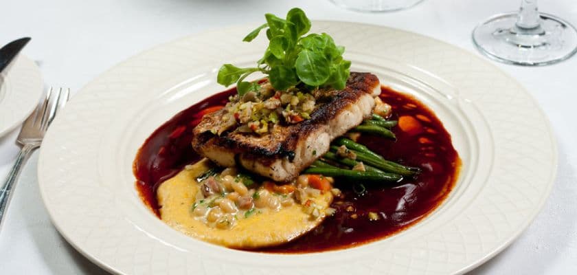 elegant plated fish entree with grilled fillet, seasonal vegetables, creamy sauce, and rich red reduction, garnished with fresh greens on a white tablecloth