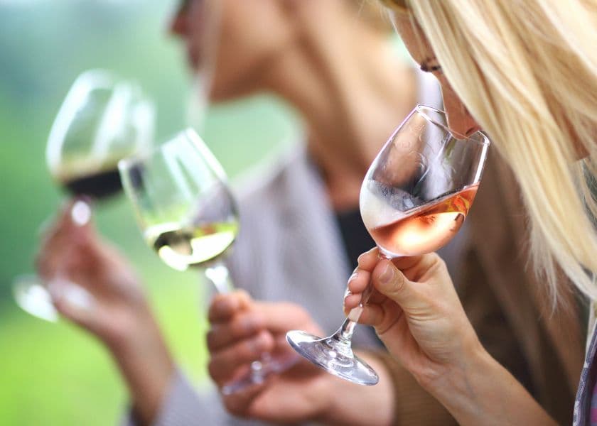 People sampling red, white, and rosé wine outdoors with soft greenery in the background.