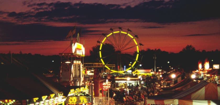 County fair at night with glowing rides, tents, and crowds.