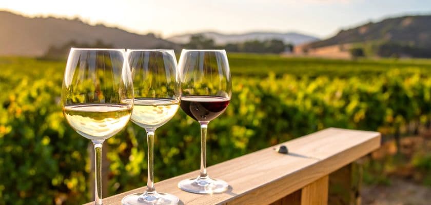 Three glasses of wine on a wooden railing overlooking a sunny vineyard and distant hills.