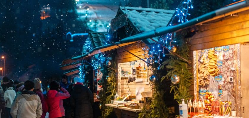 People walking through a snowy winter festival with decorated wooden vendor booths.