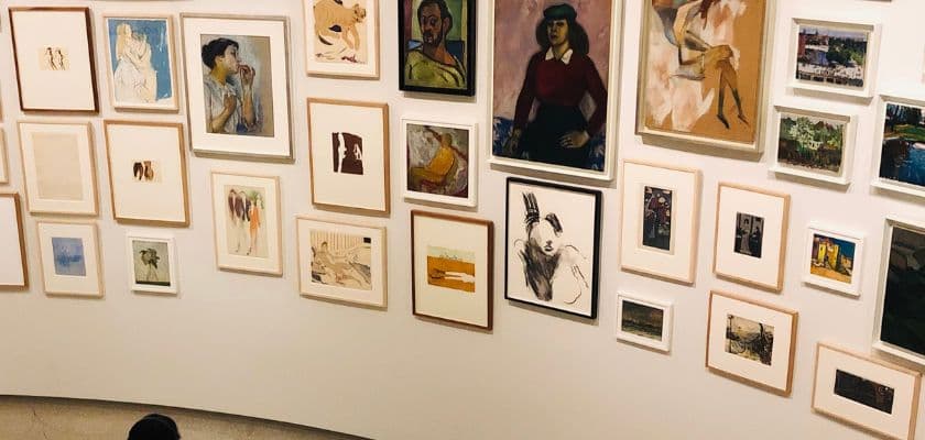 wall in art gallery displaying various framed paintings and drawings in different styles and sizes